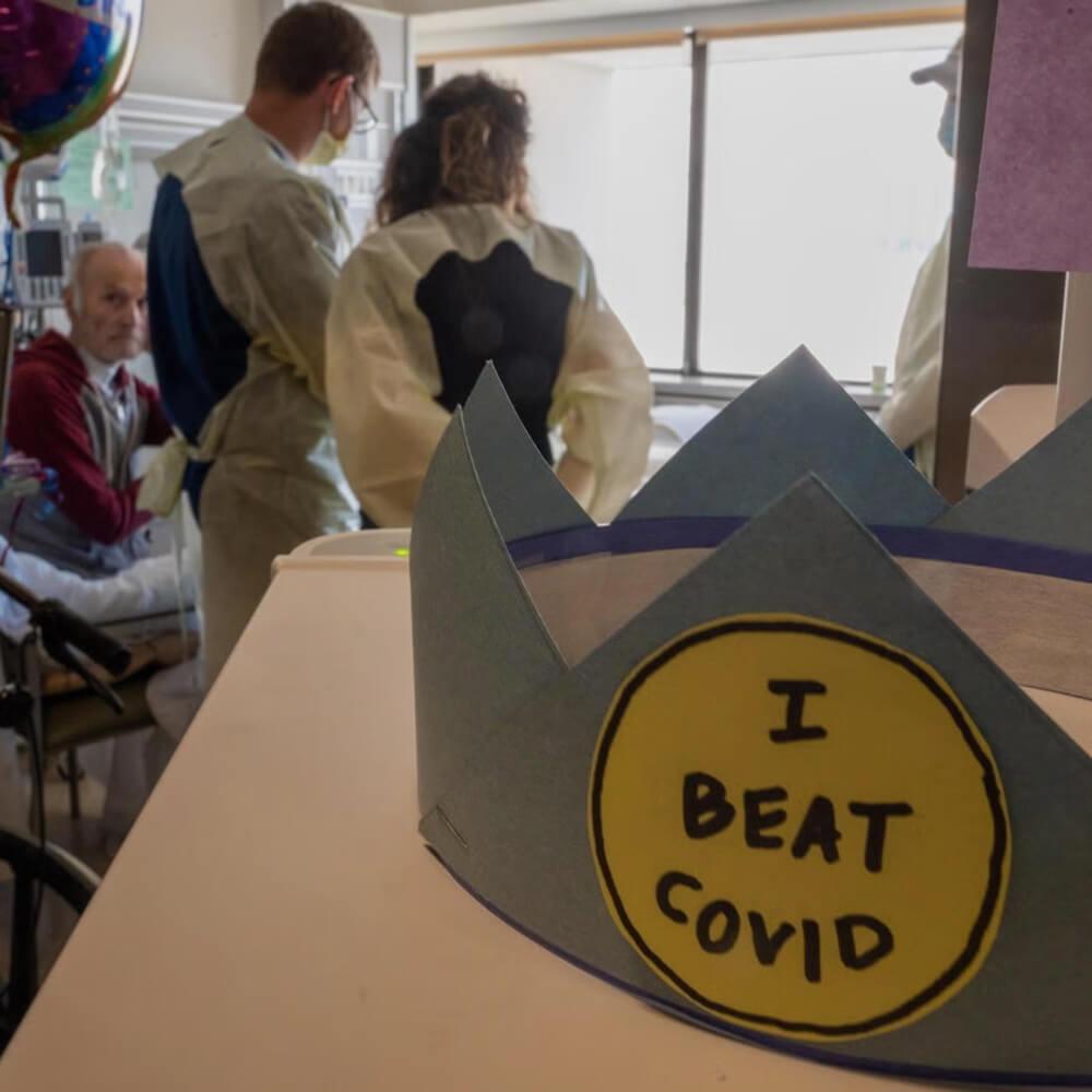 a hat in the forefront with "I beat COVID" on it while people are gathered in the background