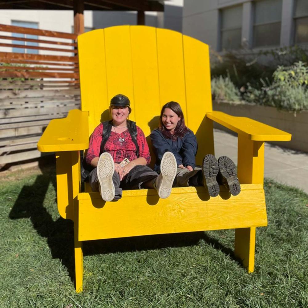 Two students sit in an oversize bright yellow Adirondack chair, smiling at the camera.
