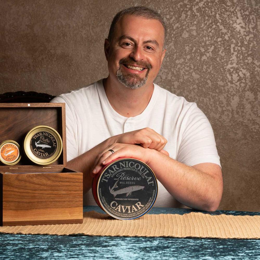 A man poses for the camera at a table behind various tins of caviar. He wears a white t-shirt and smiles.