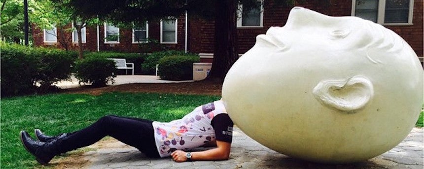 Person lying on back covering his head with an egghead statue looking up