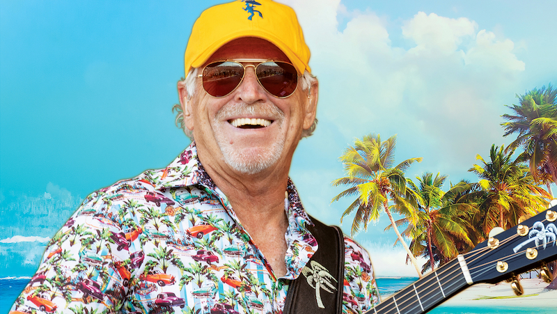 Jimmy Buffett smiling, holding his guitar, wearing sunglasses and a yellow baseball cap.