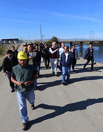 Man in yellow hardhat leads group of people down broad walkway next too river